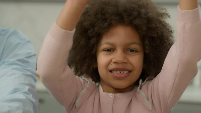 Happy carefree positive playful dancing African American ethnic child girl biracial kid daughter dance fooling having fun to music funny move at kitchen home with unrecognizable parent father smiling