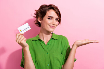 Photo portrait of attractive young woman credit card palm hold empty space dressed stylish green clothes isolated on pink color background