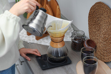 Woman pouring hot water into glass chemex coffeemaker with paper filter and coffee at countertop in kitchen, closeup