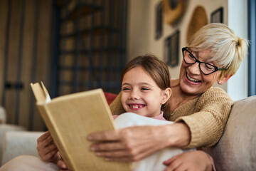 A lovely smiling grandma and her grandchild, reading a book together, sitting on the couch.