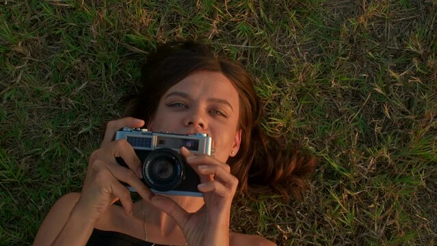 Young woman lying on the grass and takes photos with retro camera