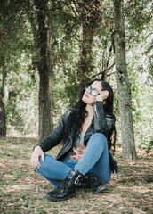 Naklejka premium Beautiful latin american brunette woman with black hair, a black jacket and blue pants sitting on the grass