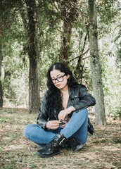 Beautiful latin american brunette woman with black hair, a black jacket and blue pants sitting on the grass