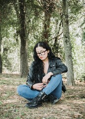 Beautiful latin american brunette woman with black hair, a black jacket and blue pants sitting on the grass