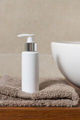 White cosmetic bottle near basin on brown folded towel on wooden countertop in bath, mockup