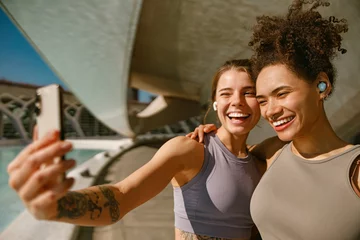 Tuinposter Two smiling female friends making selfie on a smart phone after morning run outdoors © Yaroslav Astakhov
