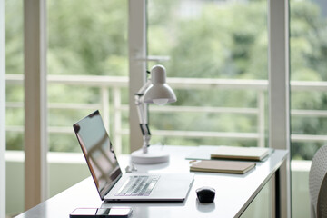 Opened laptop, planner and lamp on office desk next to big window