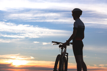 Silhouette of a cyclist next to his bike, standing still while admiring the sky and sunset in the...