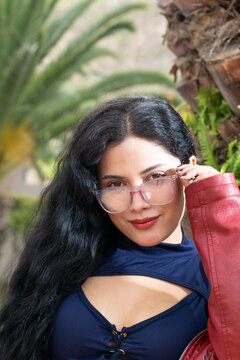 Retrato de una mujer blanca, latina, hermosa de cabello negro con chaqueta roja y una blusa azul, en frente de palmeras