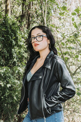 Naklejka na ściany i meble Portrait of a white beautiful latin american woman with glasses and a black jacket in the middle of the forest