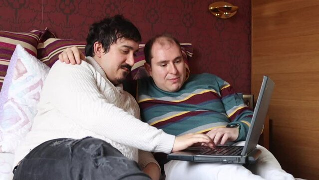 Caucasian gay couple in bed looking at laptop smiling together, side view, horizontal image with copy space. Concept of homosexual relationship