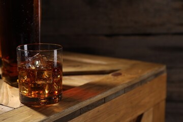 Whiskey with ice cubes in glass and bottle on wooden crate, closeup. Space for text
