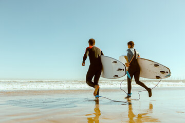 Back view of surfers with surfboards entering towards ocean for surfing on waves