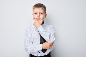 Thoughtful young boy in a shirt and vest posing with hand on chin against a gray background. - obrazy, fototapety, plakaty
