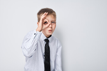 Young boy in shirt and tie making OK gesture over eye, isolated on light background.