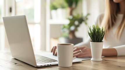 Cropped view of woman sitting at wooden table and working on laptop. A cup of coffee with a potted plant in the background. The environment is bright and clean, a relaxed work environment