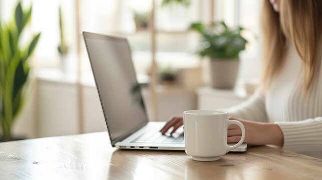 Cropped View Of Woman Sitting At Wooden Table And Working On Laptop. A Cup Of Coffee With A Potted Plant In The Background. The Environment Is Bright And Clean, A Relaxed Work Environment