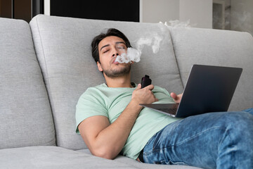 Young man using disposable electronic cigarette during work on laptop at home. Vape smoking concept