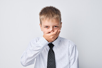 Young boy in shirt and tie covering mouth with hand, trying not to laugh, on a gray background.