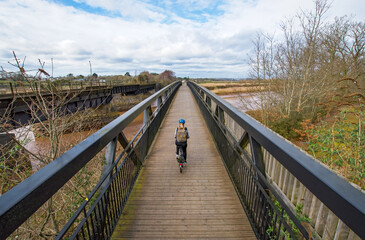 Obraz premium Cycling image of a lady on a bicycle riding across a bridge. Wide angle image with good symmetry. Selective focus on the cyclist. Concept image of a healthy lifestyle and exercise. 