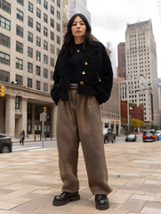 A confident urban fashion model stands on a city street, dressed in a stylish black jacket and brown trousers, embodying contemporary street style.
