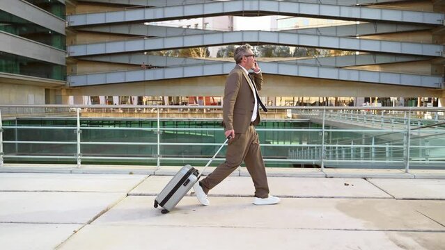 Captured in motion, this senior businessman is fully engaged on a phone call while briskly walking with his luggage, a snapshot of the dynamic nature of contemporary business, side view