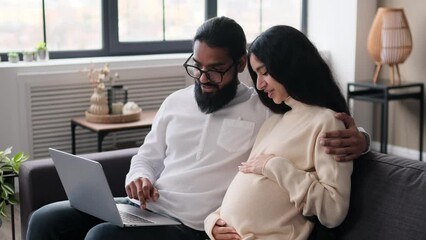 Indian husband and pregnant wife use laptop for online shopping while sitting on sofa in living room. Preparation for a child, anticipation of childbirth. - Powered by Adobe
