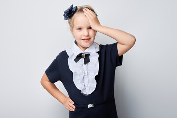 Smiling young girl in school uniform with hand on forehead against a light grey background.