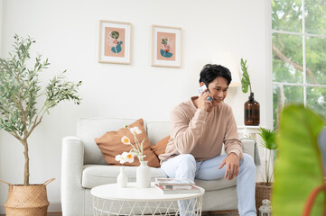 Obraz na płótnie Canvas Happy asian man sitting on couch using smartphone in living room at home, relax time and lifestyle concept