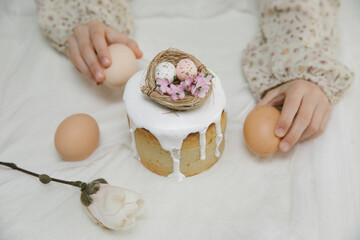 Fototapeta na wymiar Easter cake with white glaze with a decorative bird's nest with eggs and flowers in child's hands. Child is holding Easter cake. 