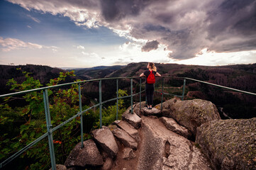 A hiker stands at a mountain lookout point, hands raised in a gesture of freedom, with a dramatic sunset sky above the vast forest landscape