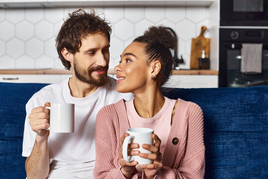 good looking jolly diverse couple in casual attire sitting on sofa and drinking coffee at home