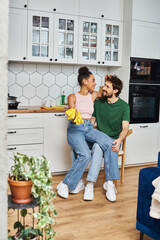 joyful beautiful diverse couple in homewear sitting on chair with rags and gloves, spring cleaning