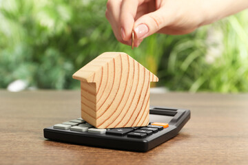 Mortgage concept. Woman putting coin into house model at wooden table against blurred green...