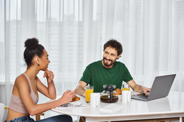 Fototapeta na wymiar joyous multiracial couple in homewear with laptop and phone smiling at each other during breakfast