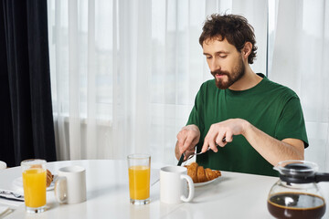 Fototapeta na wymiar handsome man in casual green t shirt cutting delicious croissant with knife during breakfast at home