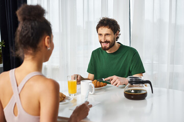 Fototapeta na wymiar joyous multiracial couple in cozy attires enjoying delicious breakfast together and smiling happily
