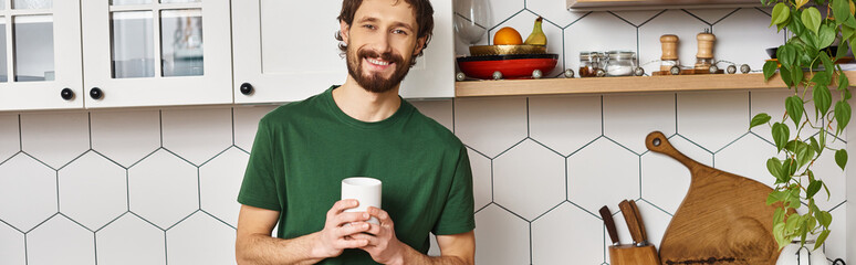 handsome jolly man in homewear holding cup with hot coffee and smiling at camera joyfully, banner