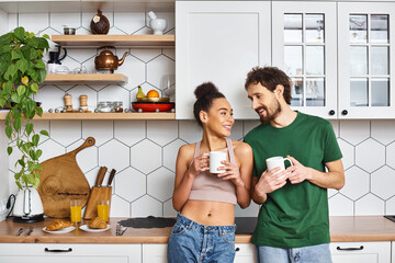 appealing cheerful diverse couple in homewear enjoying hot coffee and looking at each other