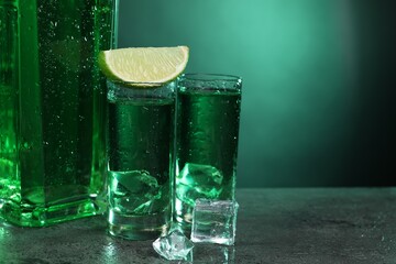 Absinthe in shot glasses, lime and ice cubes on gray textured table against green background, closeup with space for text. Alcoholic drink