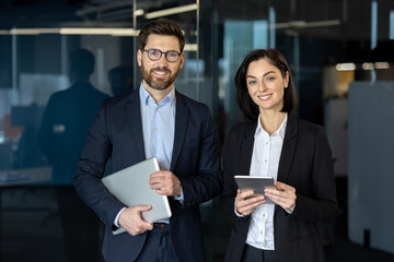 Smiling male and female in stylish suits standing together in office hall and holding digital...