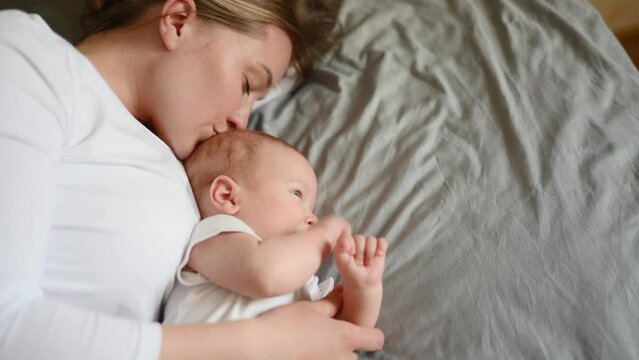 Beautiful mom and a cute little child are lying on the bed at home, the mother gently hugs the baby and kisses.Concept of motherhood, maternity leave, child care, childhood. Children's Day