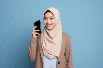 Cute young Asian girl in hijab holding a smartphone in front of a vibrant blue background