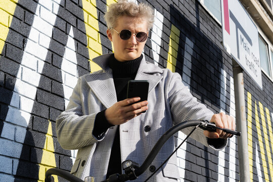 Stylish male in coat and glasses using phone while walking with retro bicycle by painted urban wall in sunny day. Neutral carbon footprint transportation. Ecofriendly mobility sustainable transport. - Powered by Adobe