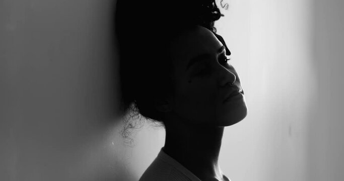 One depressed young black woman leaning on wall feeling lonely in quiet despair struggling with mental illness. Silhouette of African American 20s person suffering alone in black and white