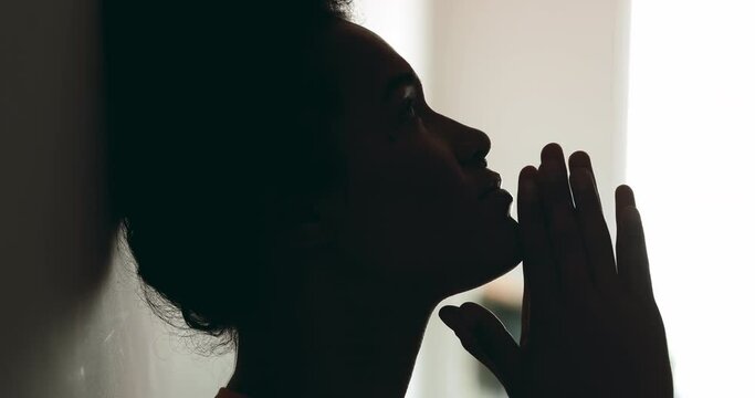One spiritual young black woman PRAYING to GOD leaning on wall in profile silhouette. Person seeking help and support during difficult times with hands clenched looking up with HOPE