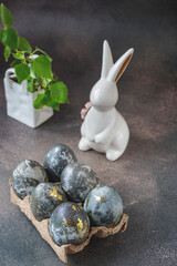 Spring Easter still life. A white rabbit and eggs painted with a gold pattern in an eco-friendly stand made of recycled cardboard on a gray background. Easter decor on the table