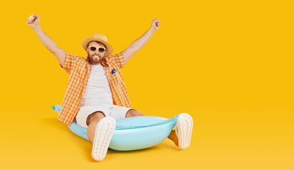 Happy smiling man in sunglasses and hat sitting on inflatable mattress with hands up isolated on a studio yellow background. Bearded guy going to swim on a beach. Vacation and holiday trip concept.