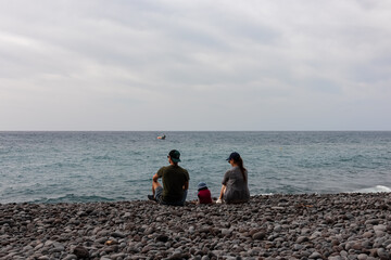 Family with small child sitting on idyllic volcanic black stone beach of Praia Garajai, Canico, Madeira island, Portugal, Europe. Waves hitting shoreline of majestic Atlantic Ocean. Vacation concept