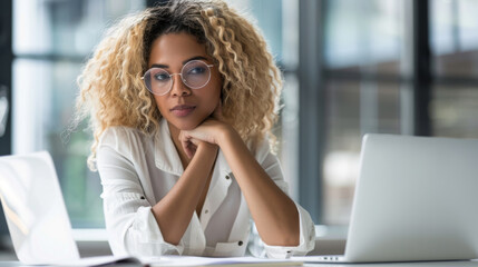 A smiling professional woman with glasses is seated in a corporate office environment with her team working in the background.